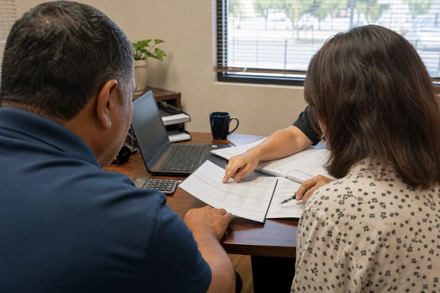 A couple seen from behind at a wooden office desk as an agent points to a column of figures on printed pages, with a laptop, calculator and window nearby